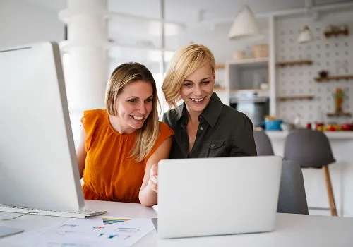 Two smiling women working together at a desk with a laptop and desktop computer, representing collaboration and digital financial solutions.