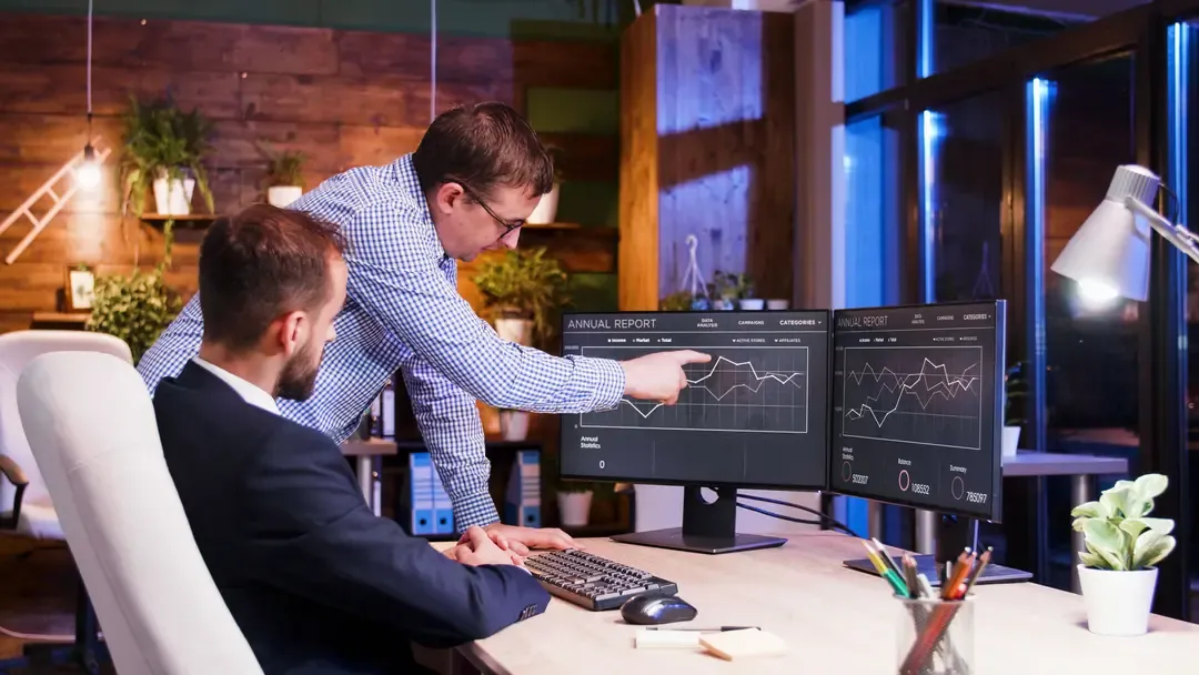 Colleague pointing at dual monitors with financial charts.
