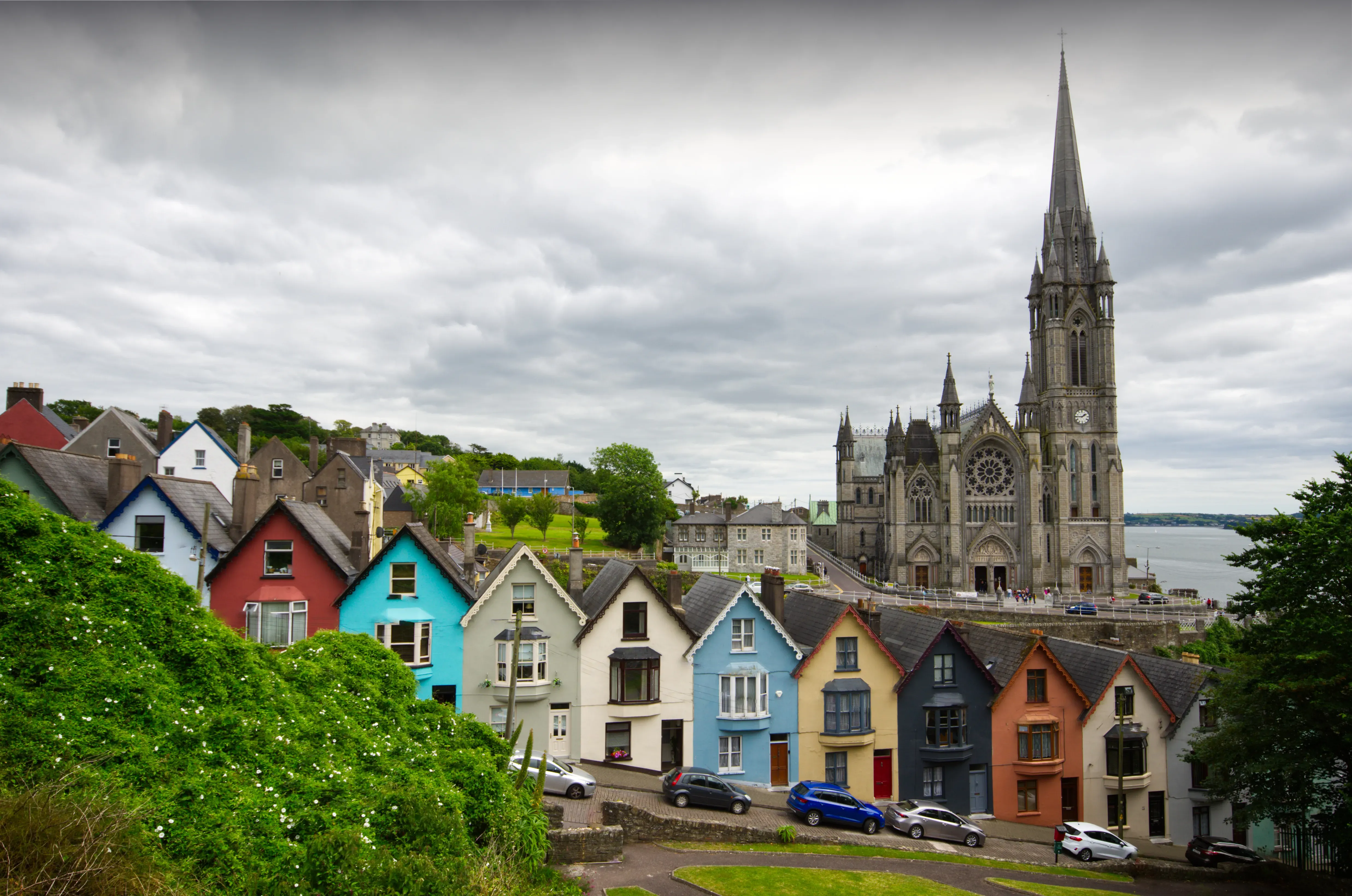 Colourful hillside houses overlooking a historic cathedral with tall spire under dramatic cloudy skies in a charming European town.