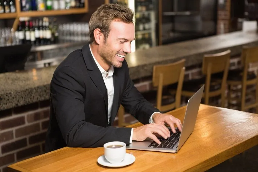 Smiling businessman working on a laptop at a coffee shop counter with a cup of coffee in front of him