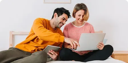 Smiling couple sitting on a couch using a tablet and laptop, representing convenient regular money transfers