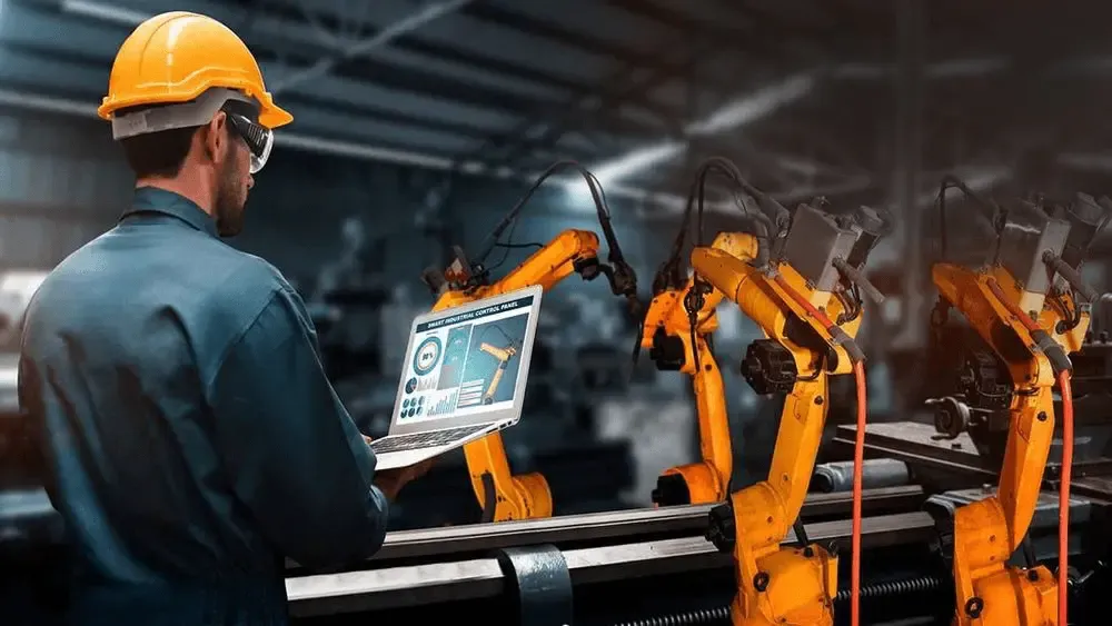 Engineer wearing a hard hat and safety glasses using a laptop to control robotic arms on an automated manufacturing production line.