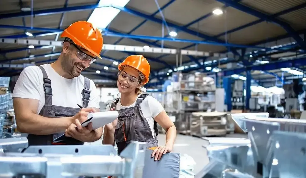 Safety hat wearing young male and female warehouse workers looking at the notepad with smiles on their faces.