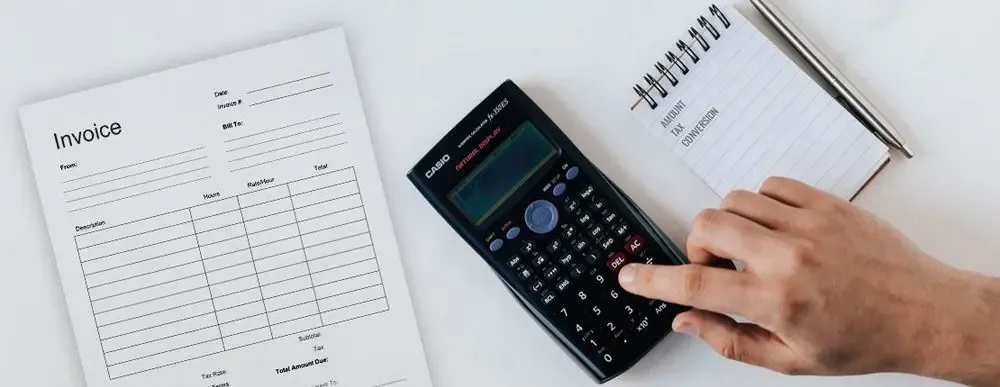 Top-down view of a hand using a calculator next to an invoice form and a notepad listing amount, tax, and conversion—symbolizing international supplier payment calculations.