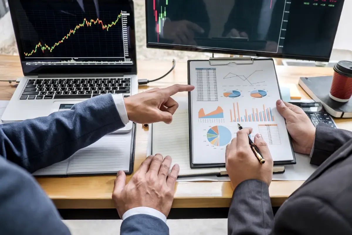 Business professionals reviewing financial charts and market data on a clipboard and computer screens while discussing currency risk and financial strategy.