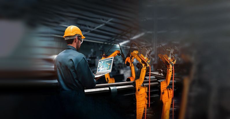 Engineer wearing a hard hat and safety glasses using a laptop to control robotic arms on an automated manufacturing production line.