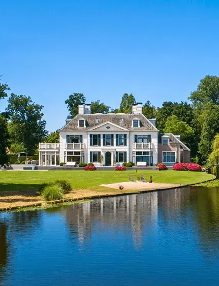Luxury waterfront property with large white house, manicured lawn, and surrounding trees under a clear blue sky.