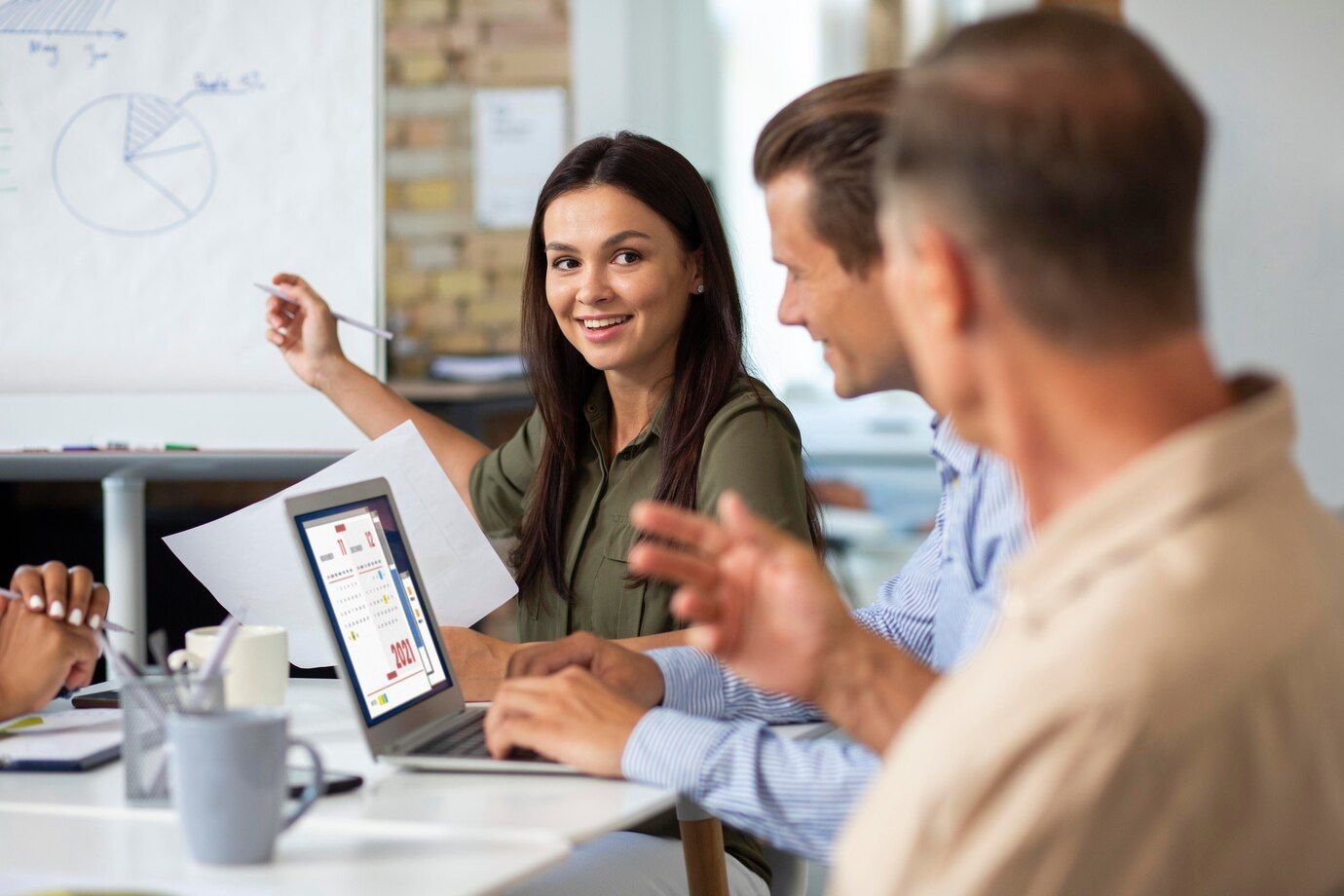 Group of professionals in a business meeting reviewing charts and calendars on a laptop, with a whiteboard graph in the background.