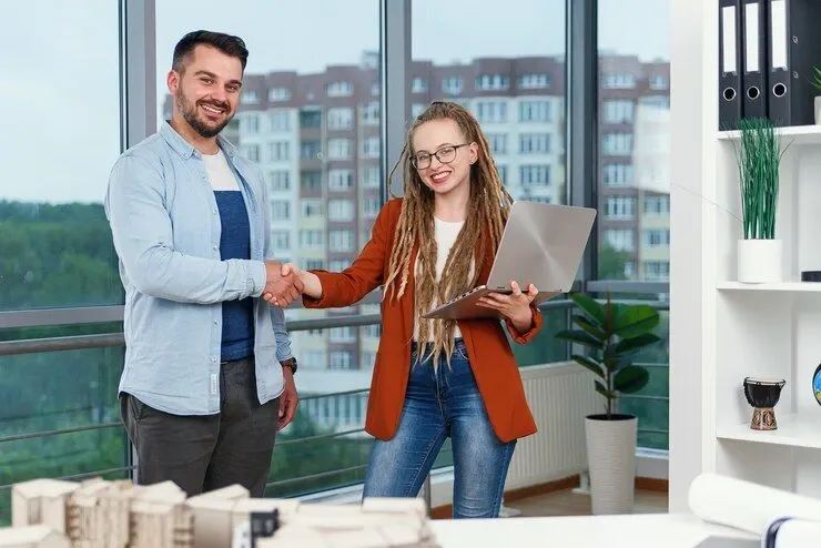 Two professionals shaking hands in a modern office with a cityscape view, holding a laptop and plans