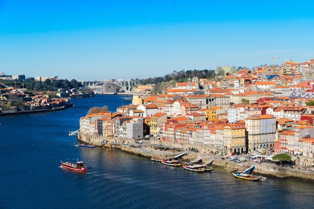 colorful riverside buildings with red-tiled roofs along the Douro River in Porto, Portugal, with boats on the water and a bridge in the distance.
