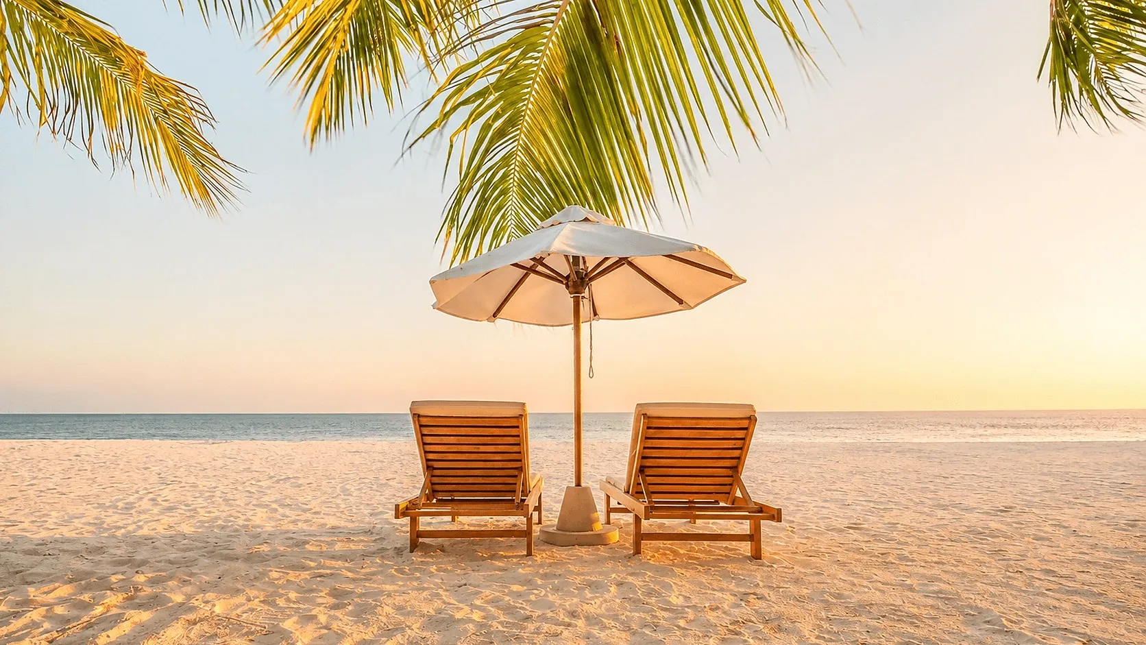 Two lounge chairs under a beach umbrella on a tropical beach at sunset, representing overseas property and vacation living.