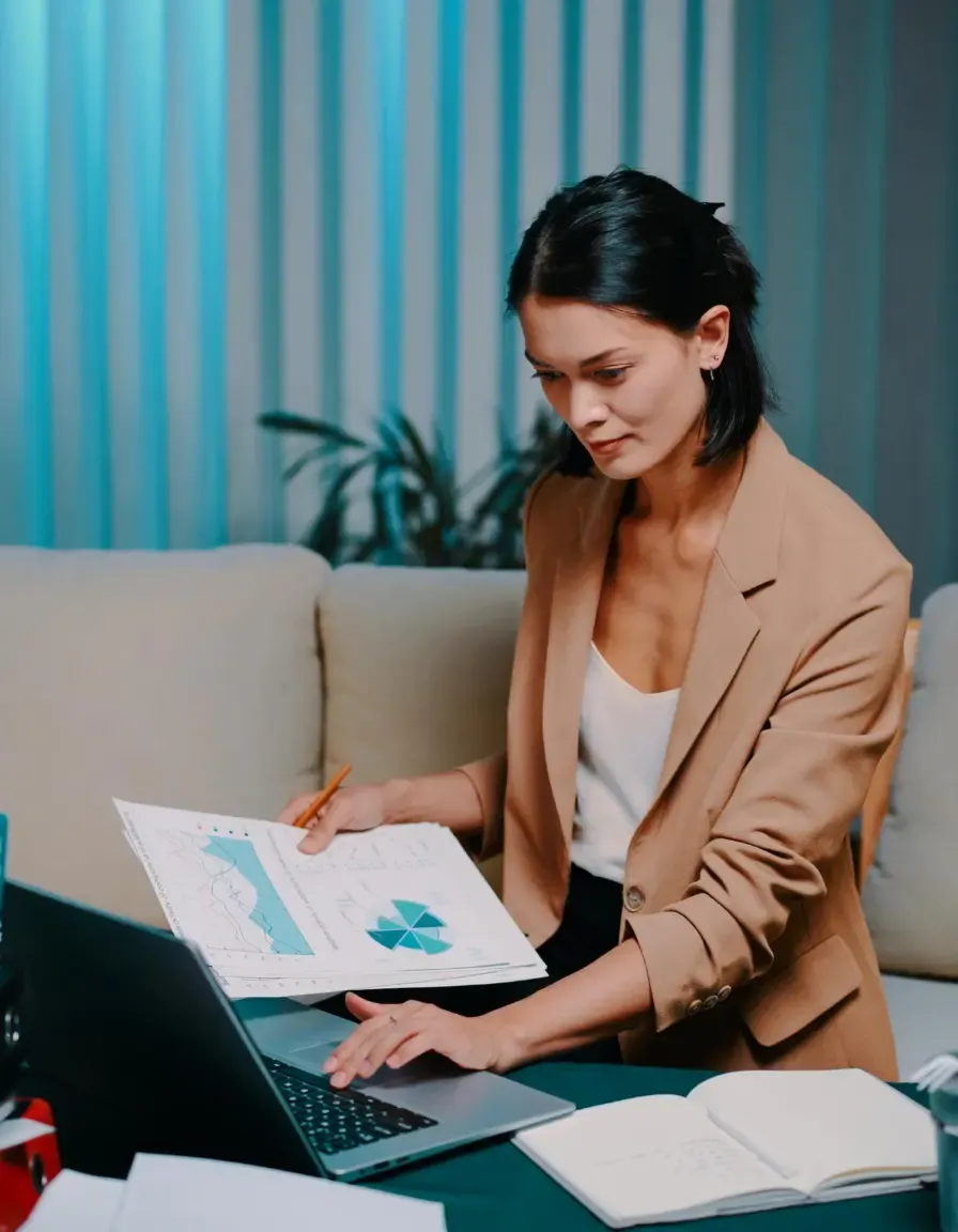 Businesswoman reviewing financial charts on paper while working on a laptop at home or office