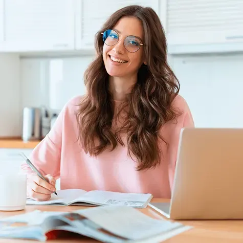 Smiling female student wearing glasses, studying with books and a laptop at a desk.