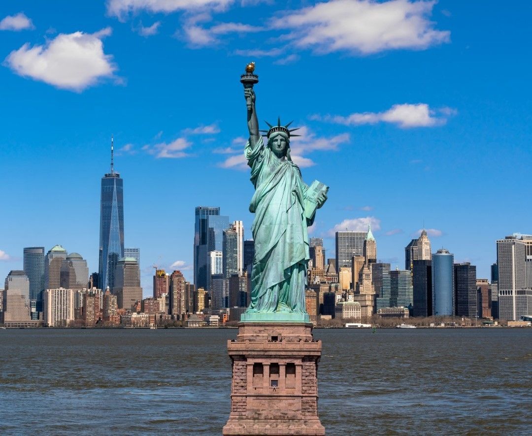 The Statue of Liberty in the foreground with the Manhattan skyline and One World Trade Center in the background on a clear, sunny day.