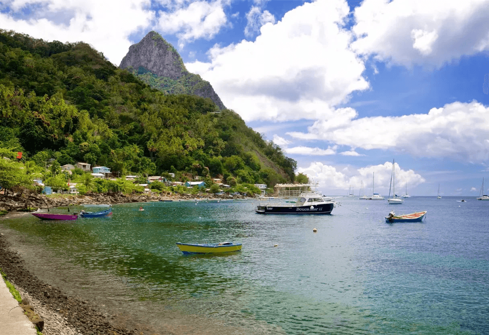 Coastal bay in St. Lucia with clear turquoise water, colourful fishing boats and yachts anchored offshore, and a lush green hillside rising to a dramatic peak under a bright blue sky, reflecting the appeal of Buying Property in St. Lucia.