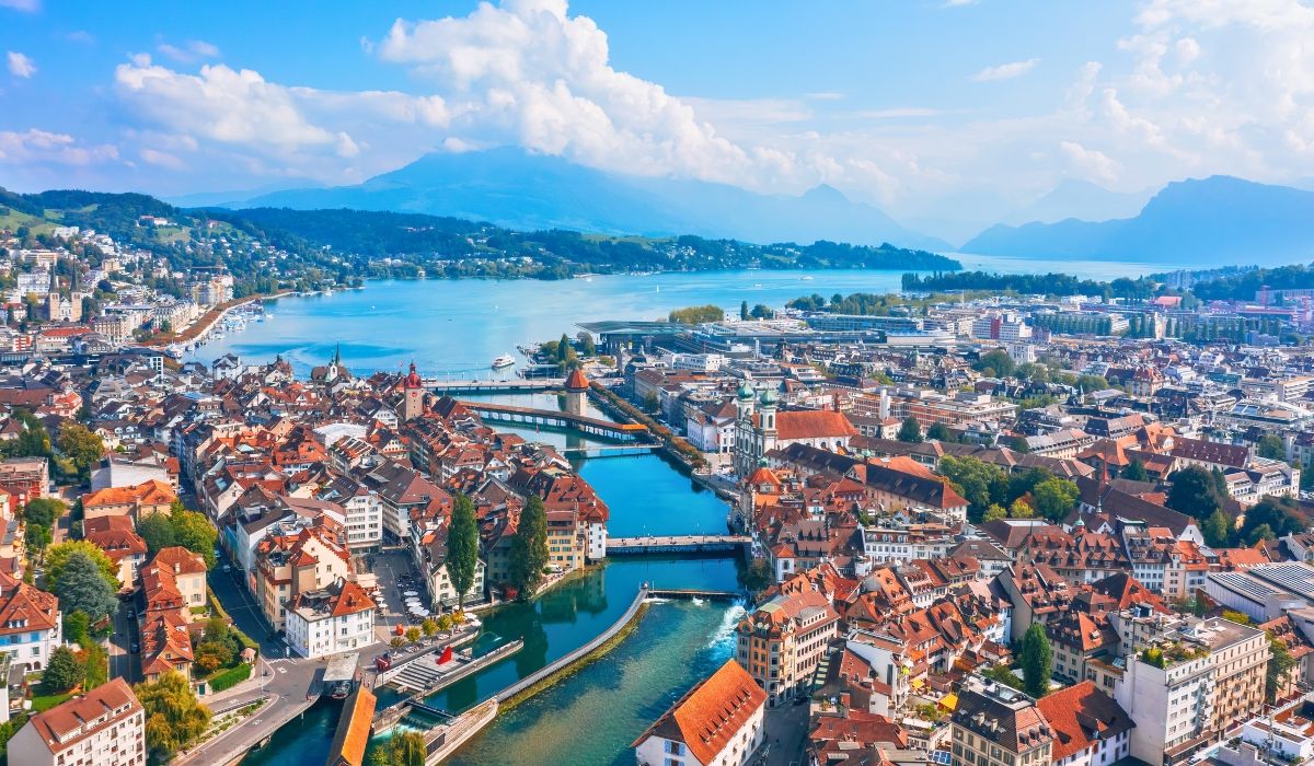 Scenic aerial view of Lucerne, Switzerland, featuring the vibrant rooftops of the old town, the Reuss River, and the picturesque Lake Lucerne, surrounded by lush green hills and distant mountains under a partly cloudy sky.