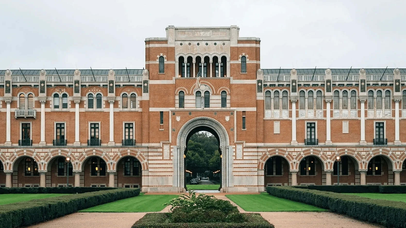 Historic university building with arched architecture, landscaped lawns, and an American flag at the entrance.