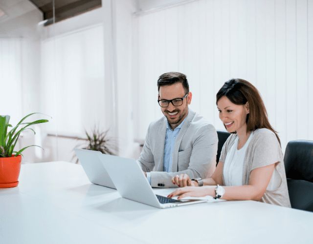Smiling man and woman working together on laptops in a bright, modern office setting.