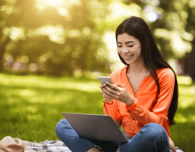 Smiling woman sitting on grass using smartphone and laptop in a sunny park