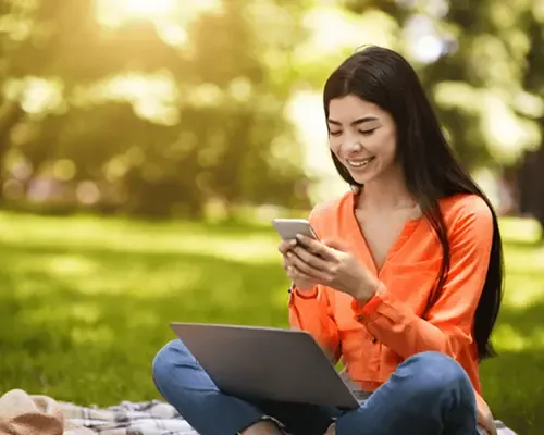Smiling woman sitting on grass using smartphone and laptop in a sunny park